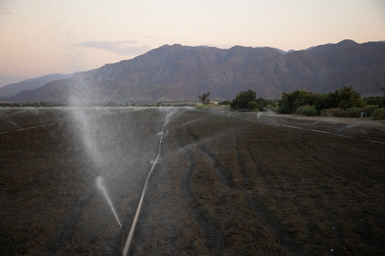 A dirt field is watered by an irrigation system, as mountains sit in the background.