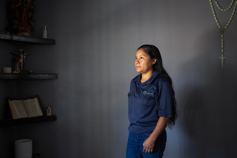 A person in a blue shirt stands near a grey wall, while a rosary hangs in the left corner and shelfs with a cross and a bible rest on the right.