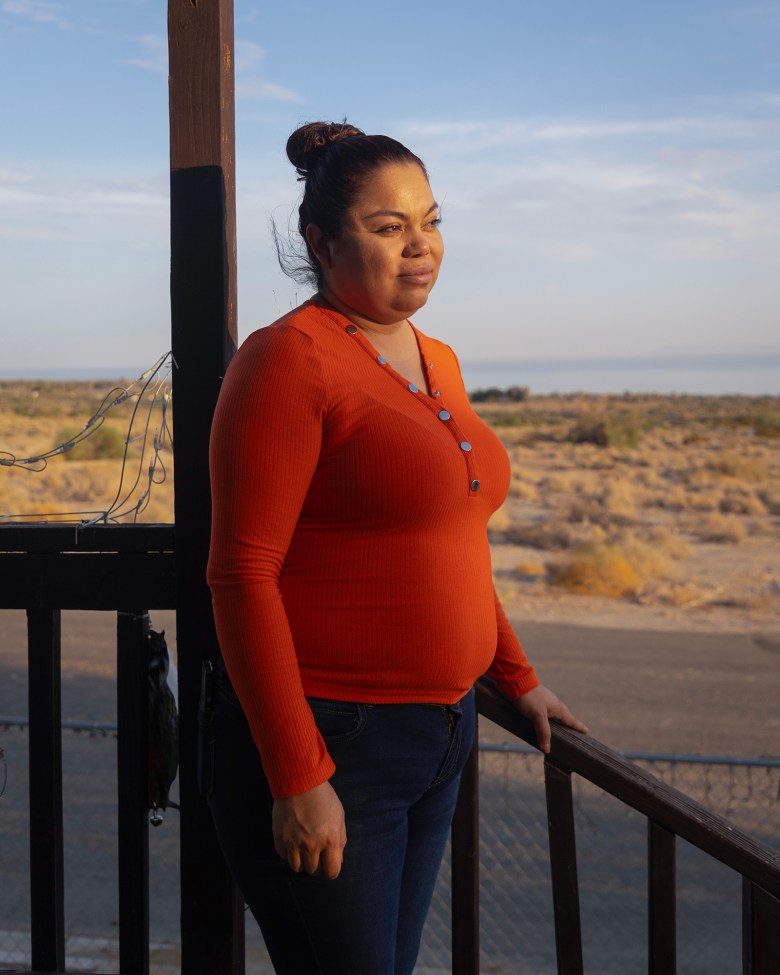 A person in an orange sweater stands on a porch facing the sun, with a desert landscape in the background.