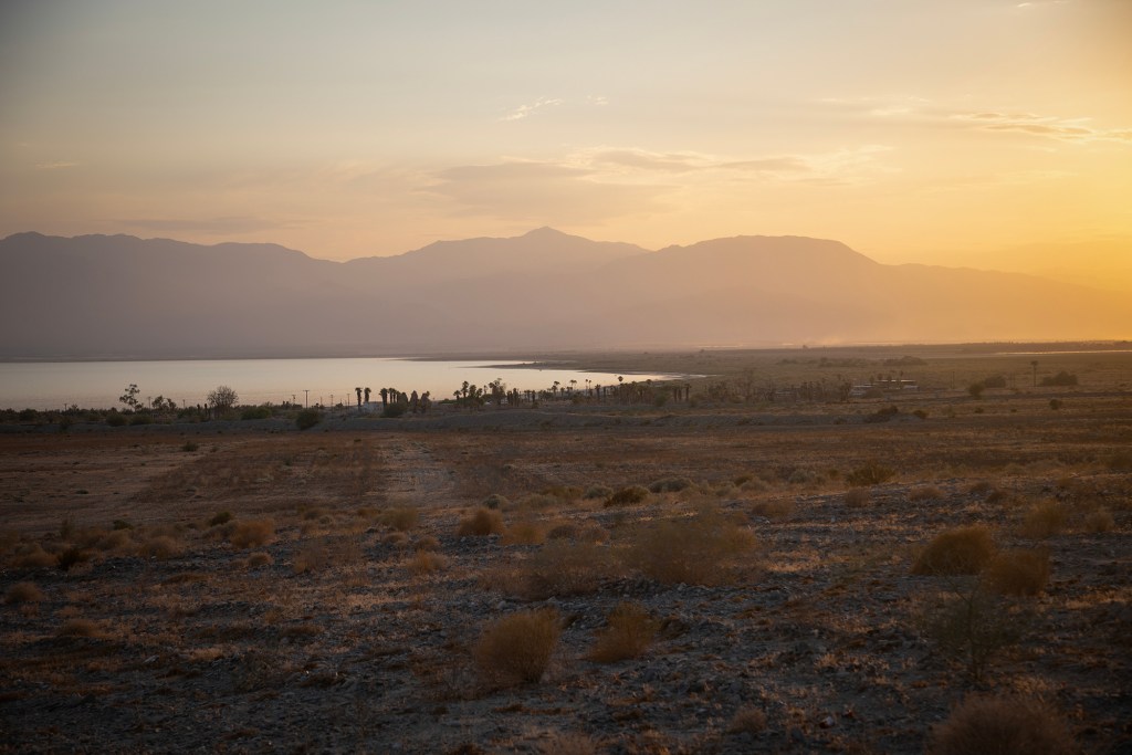 Dust particles fill the air with low sunlight filling the sky. A desert landscape with mountains in the background.